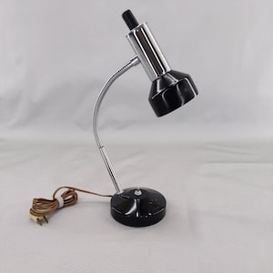 May include: A vintage desk lamp with a black shade and base, a chrome-colored flexible neck, and a black top. The lamp has a brown power cord and a two-prong plug. The lamp is designed to sit on a desk or table.