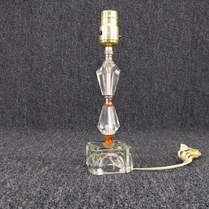 May include: A clear glass table lamp with a brass bulb socket and a decorative crystal body. The lamp has a square base and a clear electrical cord. The lamp is approximately 12 inches tall.