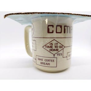 May include: A cream-colored ceramic mug with a handle, featuring a flowchart design in brown. The text on the mug includes "COMP" and "IS IT TIME TO GO HOME?" with options for "YES" and "TAKE COFFEE BREAK". The mug is packaged in cardboard.