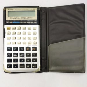 May include: A Casio FC-100 Financial Consultant calculator in a black and silver design. The calculator is open, showing its screen and many buttons. It is in a black leather-effect case with a clear plastic pocket.