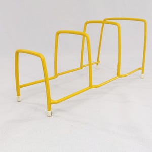 May include: A yellow metal rack with three sections, each designed to hold items. The rack has a rectangular shape with rounded corners and is supported by small white feet. The bright yellow color gives it a modern look.