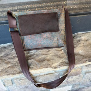 May include: A brown crossbody bag with a patterned front panel in teal, red, and gold. The bag features a dark brown leather flap and strap, with a gold-colored buckle. The strap is made of a seatbelt material.