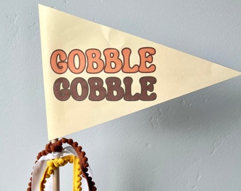 May include: A festive banner with the words "GOBBLE COBBLE" in orange and brown retro font. The banner is attached to a wooden dowel and adorned with brown and yellow pom-pom trim and white ribbons. Perfect for Thanksgiving.