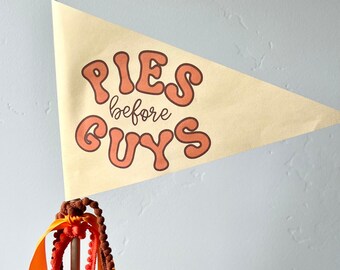 May include: A decorative pennant flag with the text "PIES before GUYS" in orange lettering. The flag is attached to a wooden dowel with orange and brown ribbons and trim.