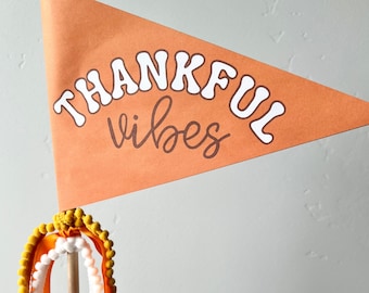 May include: Orange pennant flag with the words "THANKFUL vibes" in white and brown. The flag is attached to a wooden dowel with yellow and white pom-pom trim and orange ribbons. A festive decoration.