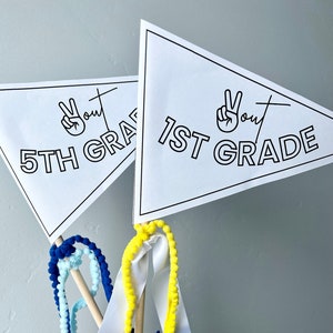May include: Two white triangular pennants with black text and a peace sign graphic. The pennants read "1st Grade" and "5th Grade" with the word "out" written next to the peace sign. The pennants are attached to wooden sticks with colorful pom-pom streamers.