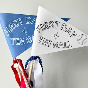 Könnte beinhalten: Zwei blaue und weiße Wimpel mit dem Text "First Day of Tee Ball" und einer Baseballgrafik. Die Wimpel sind an Holzstöcken mit roten und weißen Bändern befestigt.