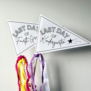 May include: Two white triangle pennants with black text and a black star. The top pennant reads "Last Day of Kindergarten". The bottom pennant reads "Last Day of Fourth Grade". The pennants are attached to wooden sticks with colorful pom-pom ribbon streamers.