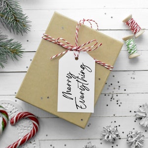 May include: A wrapped gift box with a white tag that reads "Merry Everything". The box is tied with red and white twine. Candy canes, pine needles, and star-shaped confetti are scattered around the box on a white wooden surface. Wooden spools of red and green twine are also present.