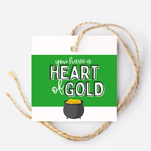 Op de afbeelding: Een wit cadeaulabel met een groene streep en de tekst "you have a HEART of GOLD". Een zwarte pot met gouden munten staat onderaan. Het label is vastgebonden met natuurlijk touw.