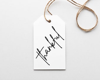 May include: A white tag with the word "Thankful" in black script. The tag is suspended by a brown string, set against a white background. The design is simple and elegant.