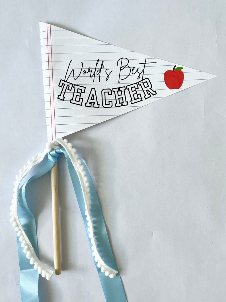 K&ouml;nnte beinhalten: Ein wei&szlig;es Wimpel mit roten Linien und dem Text "World's Best Teacher" in Schwarz. Eine rote Apfelgrafik befindet sich auf der rechten Seite des Wimpels. Der Wimpel ist an einem Holzstab mit einem blauen Band mit wei&szlig;en Pompons befestigt.