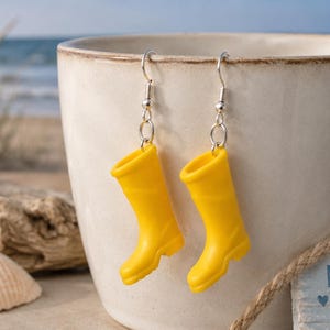 Könnte beinhalten: Ein Paar gelbe Gummistiefel-Ohrringe. Die Ohrringe hängen an silbernen Haken. Sie sind vor einem weißen Becher platziert, mit einer Strandkulisse im Hintergrund.