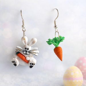 Könnte beinhalten: Ohrringe mit Ostermotiv. Ein Ohrring zeigt einen weißen Hasen, der eine orangefarbene Karotte hält. Der andere Ohrring ist eine orangefarbene Karotte mit grünen Blättern. Beide haben silberfarbene Ohrhaken.