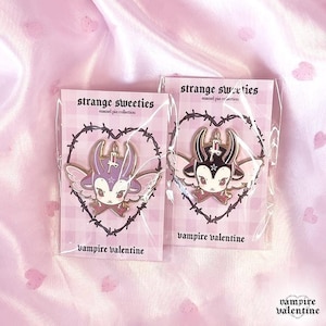 May include: Two enamel pins from the strange sweeties collection, each featuring a winged creature with horns and a candle, displayed on backing cards with a barbed wire heart design and the text strange sweeties and vampire valentine.