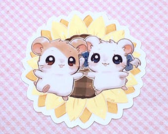 Könnte beinhalten: Ein gestanzter Aufkleber mit einem hellbraunen Hamster und einem weißen Hamster mit blauer Schleife, beide mit großen dunklen Augen, vor einer Illustration einer Sonnenblume mit dunkelbrauner Mitte und hellgelben Blütenblättern.