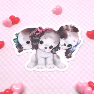 Puede incluir: Una pegatina que muestra tres cachorros ilustrados con grandes ojos oscuros. El cachorro central lleva un lazo rosa, el de la izquierda tiene un lazo verde y el de la derecha tiene un lazo azul. Los cachorros tienen pelaje blanco y marrón.