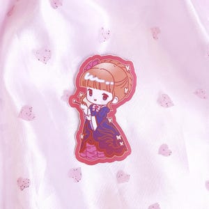 May include: A sticker featuring a cartoon girl with brown hair, wearing a purple and pink dress with butterfly accents. The sticker has a pink background.