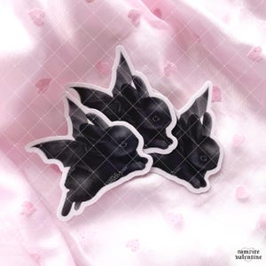 May include: Three die-cut stickers featuring black rabbits with bat wings. Each sticker shows a dark-furred bunny with pointed ears and leathery wings extended. The stickers have a white border and include the text SAMPLE printed on the surface.