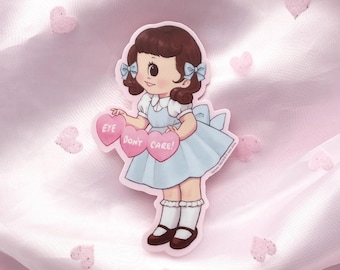 May include: A die-cut sticker featuring a cartoon character with brown hair, blue bows, and a light blue dress. The character holds three pink heart-shaped signs that read EYE, DON'T, and CARE! The sticker has a white border and a matte finish.