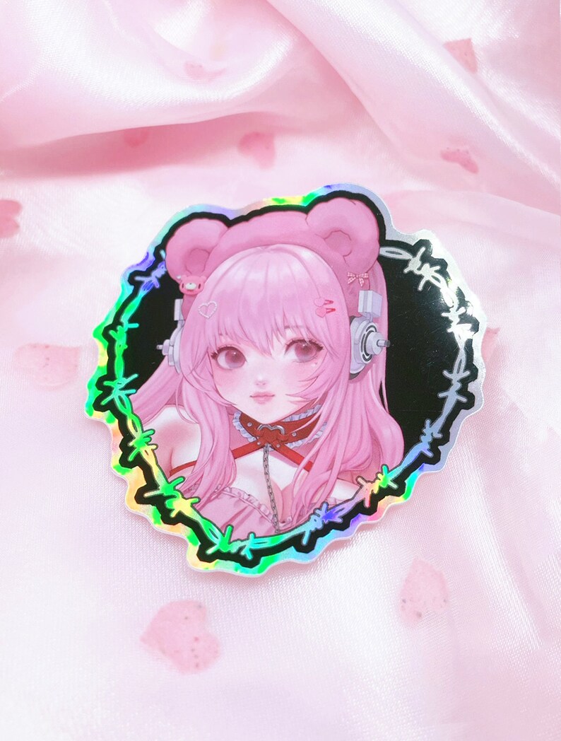 May include: A holographic sticker featuring a pink-haired anime character with bear ears, wearing a choker and headphones. The character is surrounded by a black barbed wire border.