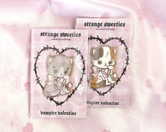 May include: Two enamel pins on backing cards labeled strange sweeties enamel pin collection and vampire valentine. One pin features a grey kitten with a pink bow, the other a calico kitten with a pink bow, both framed by a black barbed wire heart.