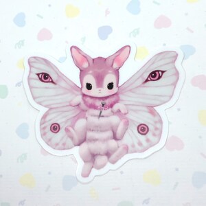 May include: A pink and white moth with large eyes and a fuzzy body. The moth has a pink rabbit head and is holding a pink flower.