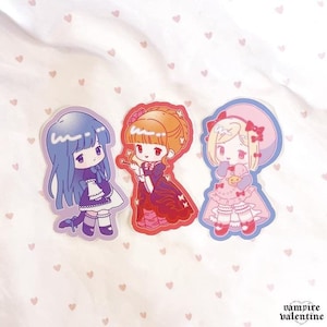 Könnte beinhalten: Drei gestanzte Aufkleber mit Charakteren im Chibi-Stil. Die linke Figur hat blaues Haar und ein violettes Kleid. Die mittlere Figur hat orangefarbenes Haar und ein rotes Kleid. Die rechte Figur hat blondes Haar, ein rosa Kleid und einen Hut.
