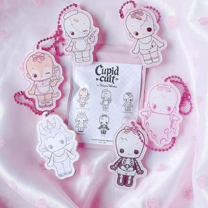 May include: Five acrylic keychains with pink ball chains featuring illustrated characters including a nurse, a ghost, and a skeleton, arranged around a white paper packet labelled Cupid Cult by Vampiie Valentine with small character icons printed on it.
