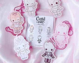 May include: Five acrylic keychains with pink ball chains featuring illustrated characters including a nurse, a ghost, and a skeleton, arranged around a white paper packet labeled Cupid Cult by Vampiie Valentine with small character icons printed on it.