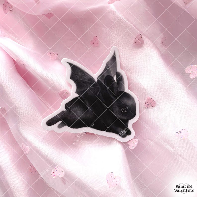 May include: A die-cut sticker featuring a black rabbit with large, dark bat wings. The rabbit is depicted in a side profile, showing its ears and fur texture. The sticker has a white border and is placed against a soft, light pink fabric background.