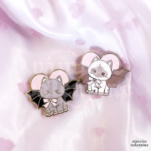 May include: Two enamel pins featuring cartoon cats with bat wings and pink heart backgrounds. One cat is grey with black wings, the other is white with brown wings. Both cats wear light pink bows around their necks. The text vampire valentine is visible.