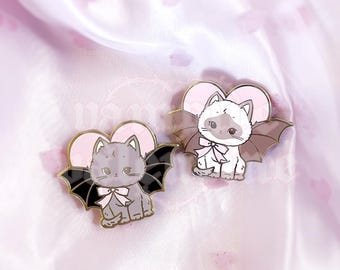 May include: Two enamel pins featuring cartoon cats with bat wings and pink heart backgrounds. One cat is grey with black wings, the other is white with brown wings. Both cats wear light pink bows around their necks. The text vampire valentine is visible.