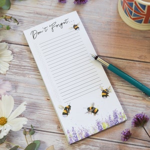 May include: A white notepad with the text "Don't Forget..." and a line for writing. The notepad has a floral design with bees on it. A teal pen is lying next to the notepad.