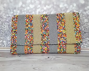May include: A rectangular clutch with a flap closure. The bag features vertical stripes of gray, gold, and colorful beaded accents. The beads are in various colors, including red, yellow, blue, and green. The background is a blurred silver.