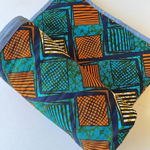 May include: A folded, patterned fabric with a denim blue border. The fabric features a repeating geometric design in shades of orange, teal, and navy blue. The pattern includes squares and striped elements, creating a vibrant and textured appearance.