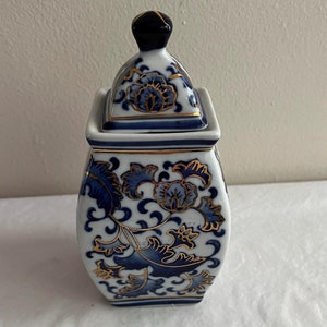 May include: A white and blue porcelain jar with a lid. The jar features a floral design in blue and gold. The lid has a black knob. The jar is a decorative item.