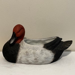 May include: A ceramic planter shaped like a duck with a red head, black and white body, and a white interior.