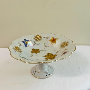 May include: A white, footed serving dish with a scalloped edge and gold trim. The dish is decorated with gold and blue leaf and floral designs. The pedestal base has decorative cutouts.