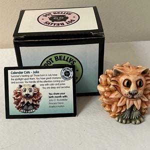 May include: A ceramic figurine of a cat with a lion mane, in a light brown color. The figurine is part of the "Pot Belly's" collection and is labeled "Calendar Cats - Julia". The text on the card reads: "Summer's heating up! Those born in July have the spotlight upon them. You have great charisma and success. You handle all the attention coming your way with calm and poise. You are deep and secretive. You share your birth month with: John D. Rockefeller, Princess Diana, Anjelica Huston."