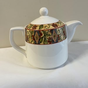 May include: White ceramic teapot with a decorative band featuring floral and geometric patterns in red, green, and gold. The teapot has a lid with a round knob and a curved spout. The handle is a square shape.