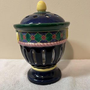 May include: A ceramic incense burner with a navy blue base and body, featuring a decorative green, yellow, and red diamond pattern. The lid is navy blue with small holes and a yellow finial. The burner has a decorative pink rope design.