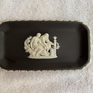 May include: A rectangular black tray with a cream-colored border. The tray features a white relief of two figures in classical attire. The border has a repeating pattern. The tray is likely made of ceramic or a similar material.