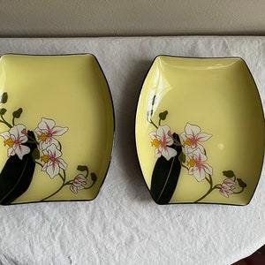 May include: Two yellow ceramic serving dishes with black rims. Each dish features a floral design of white orchids with pink accents on a black stem.