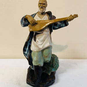 May include: A ceramic figurine of a man playing a stringed instrument. The figure wears a white tunic, green shorts, and a dark blue coat with a patterned trim. The man has a white beard and is standing on a dark base.