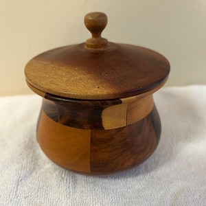 May include: A small, round wooden box with a lid. The box is made of various shades of brown wood, with a darker wood used for the lid and a lighter wood for the body. The lid has a small, rounded knob. The box is sitting on a white surface.