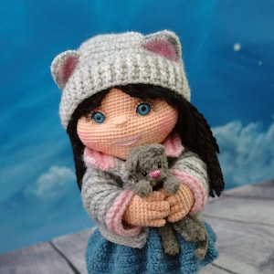 May include: A crocheted doll wearing a gray hat with pink ears, a pink and gray sweater, a blue skirt, and pink and gray boots. The doll is holding a gray crocheted cat.