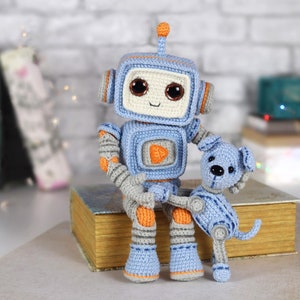 Könnte beinhalten: Ein blaues und oranges Häkelroboter-Spielzeug, das ein blaues Häkelhund-Spielzeug hält. Der Roboter hat große braune Augen und ein freundliches Lächeln. Die Spielzeuge sitzen auf einem Stapel Bücher.