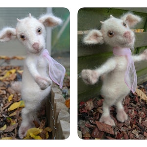 May include: A white crocheted goat doll with a pink scarf standing on a wooden fence. The goat has a white body, brown eyes, and a pink nose. The goat is standing on a wooden fence with brown leaves on the ground.