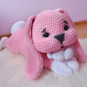 Puede incluir: Un juguete de peluche de conejo rosa tejido a crochet con pies y orejas largas de crochet blanco. El conejo tiene ojos negros y una nariz negra.